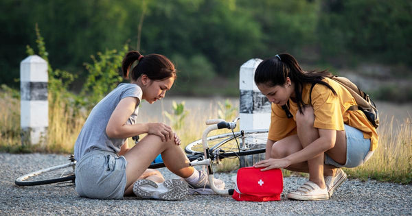 A girl helping another girl who fell off a bike using a first aid kit